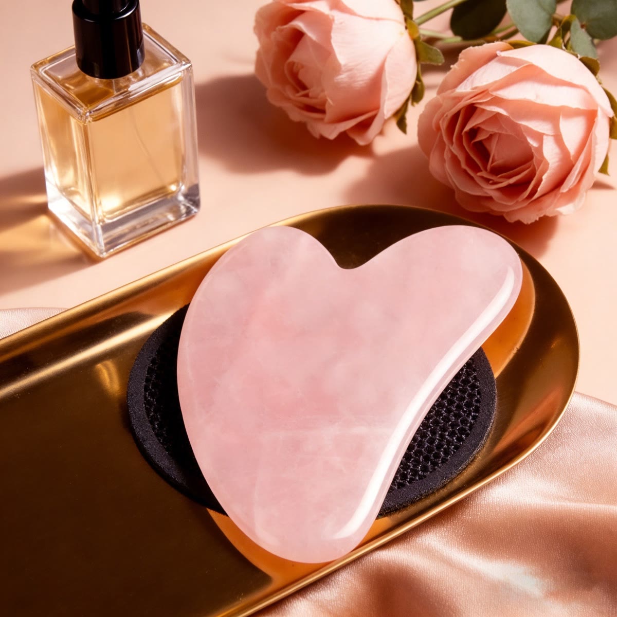 Gua sha i rosenkvarts på et guldfad ved siden af roser og en parfumeflaske