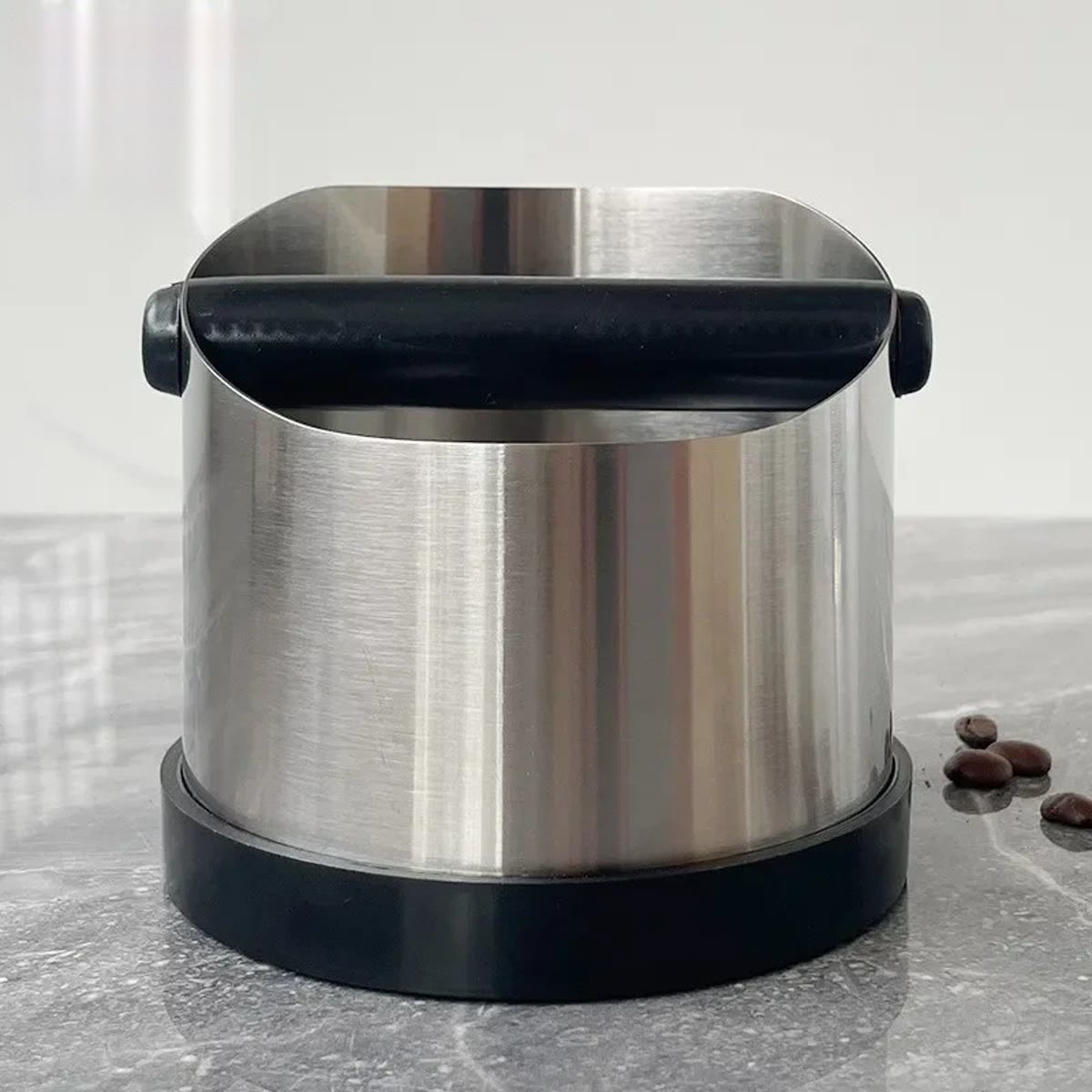 Knock boxen set forfra på en grå marmorbordplade med stangen på tværs af åbningen og et par kaffebønner ved siden af