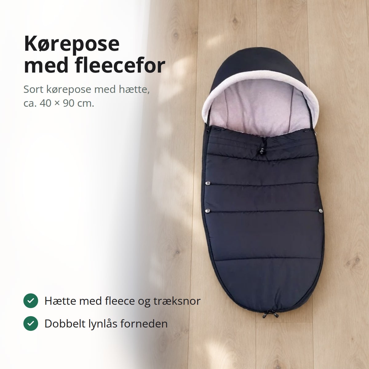 Sort kørepose med lyst fleecefor og hætte, liggende fladt på et lyst egetræsgulv ved et vindue