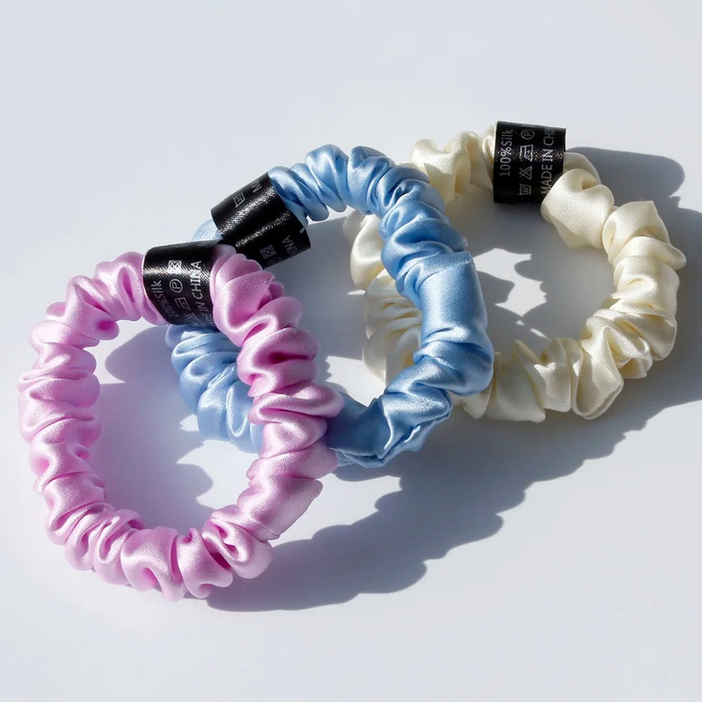 Tre silke scrunchies i rosa-lilla, lyseblå og creme med sort etiket i sollys
