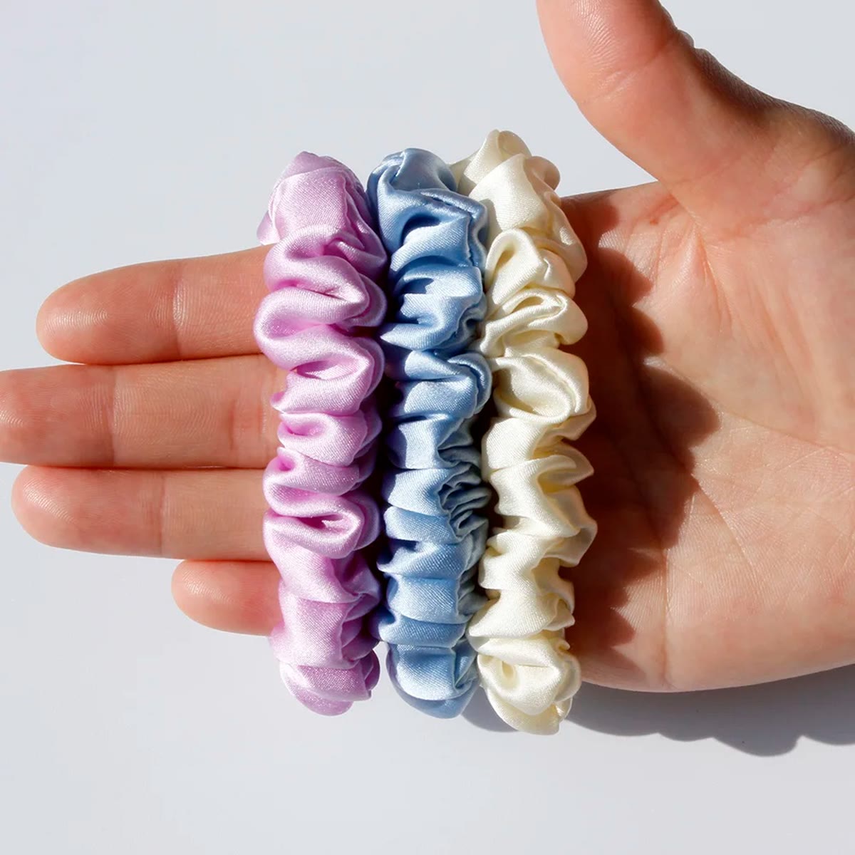 Hånd, der holder tre smalle silke scrunchies i rosa-lilla, lyseblå og creme