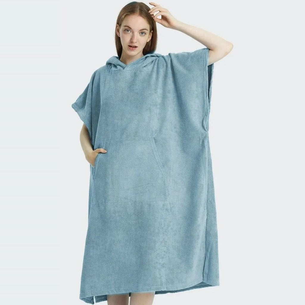 Kvinde i lyseblå badeponcho med hætten nede og hånden i kængurulommen, set forfra i fuld længde mod en lysegrå baggrund