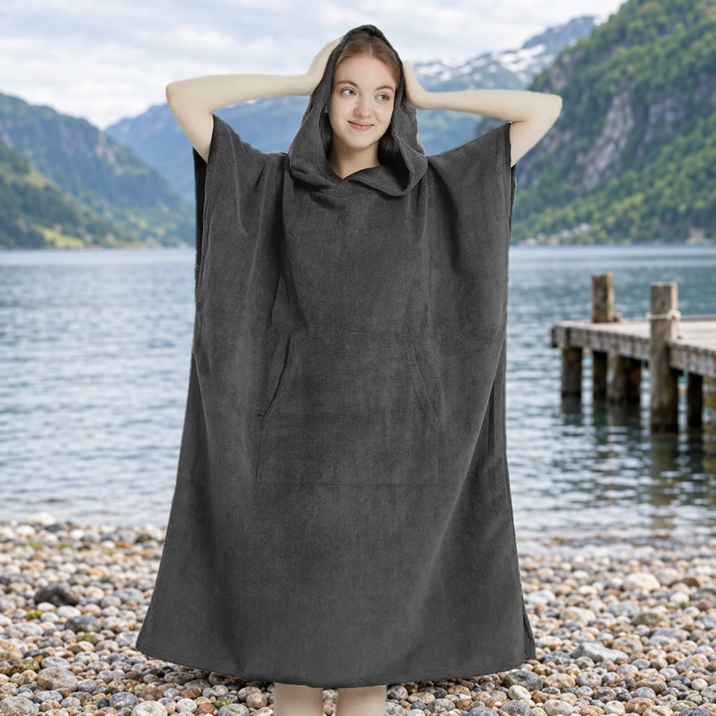 Kvinde i mørkegrå badeponcho med hætten oppe på en stenstrand ved vandet, med en badebro og bjerge i baggrunden