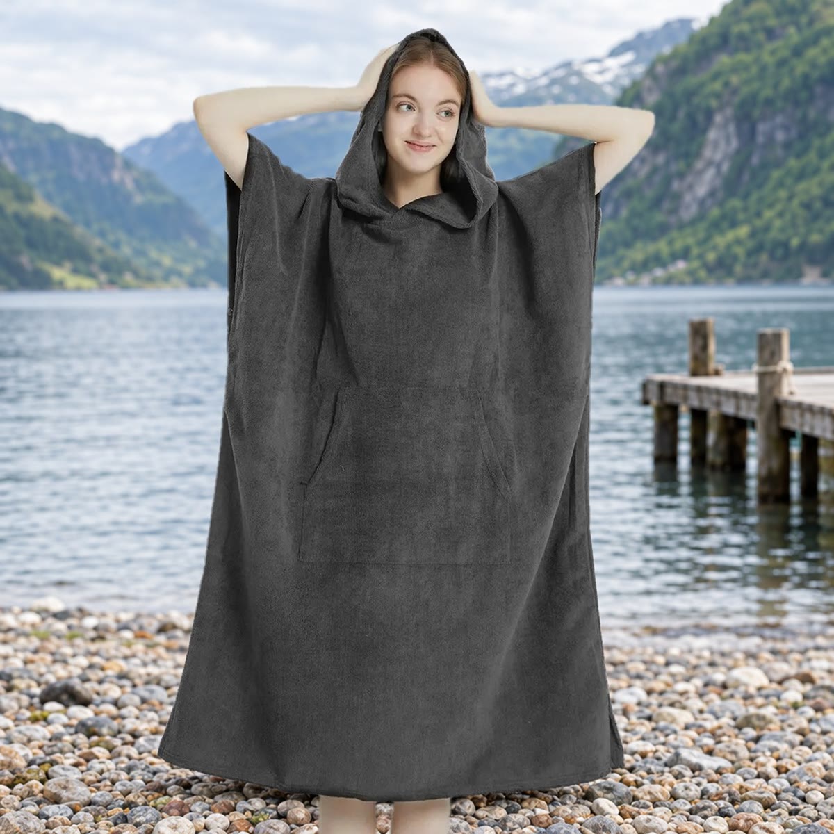 Kvinde i mørkegrå badeponcho med hætten oppe på en stenstrand ved vandet, med en badebro og bjerge i baggrunden