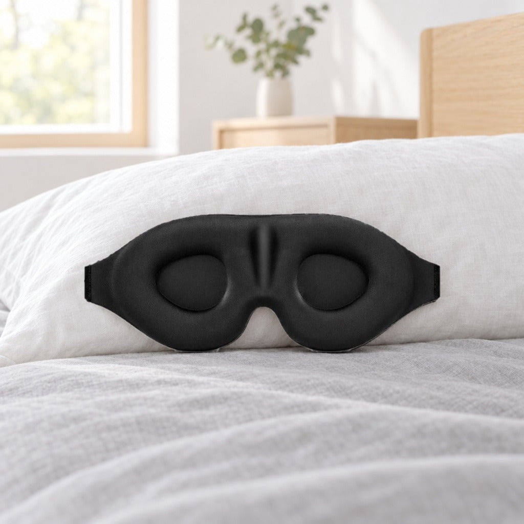 Sort 3D-sovemaske med ovale øjenkopper og formet næseryg, lænet op ad en hvid pude på en seng