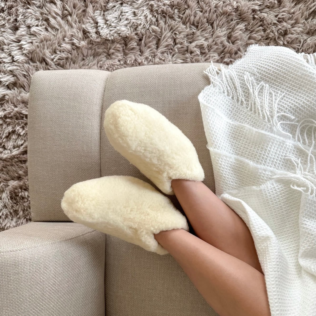 Uldhjemmesko i hvid uld på en sofa med et plaid over benene