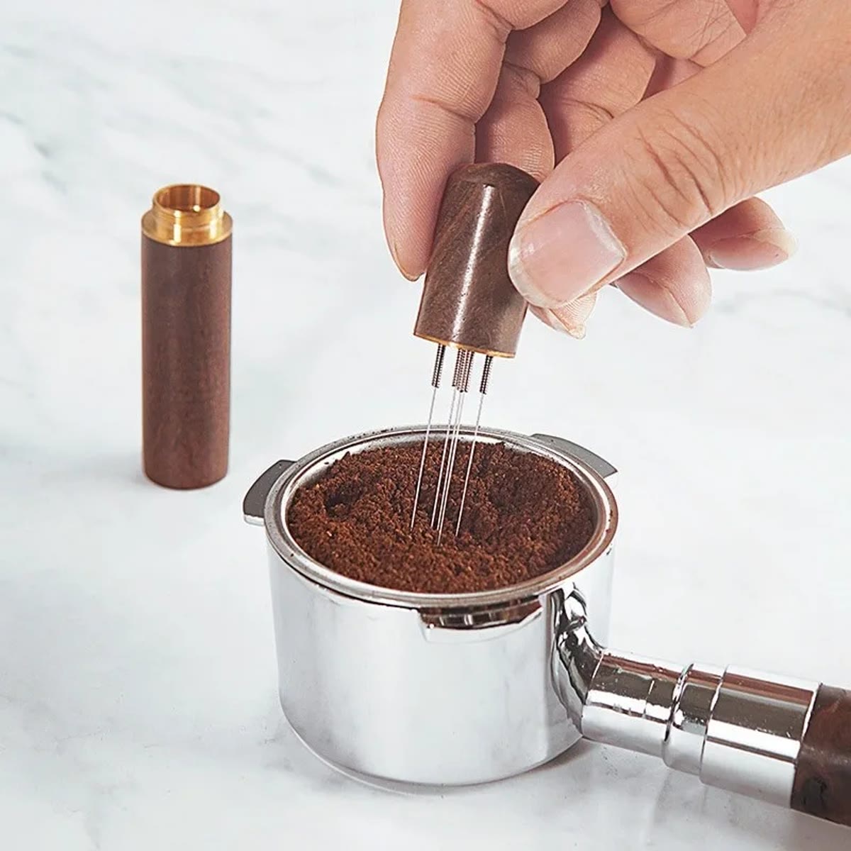 Hånd, der rører med WDT-værktøjet i den malede kaffe i en filterkurv, med låget ved siden af
